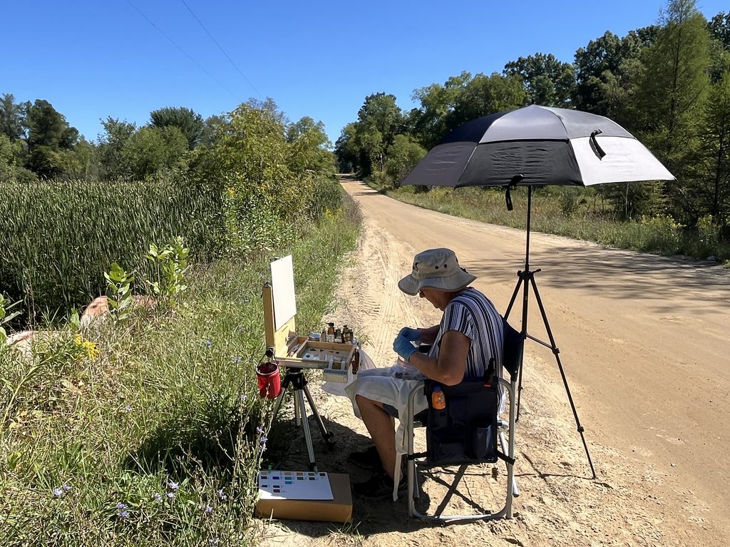 Eileen sitting under an umbrella painting outdoors